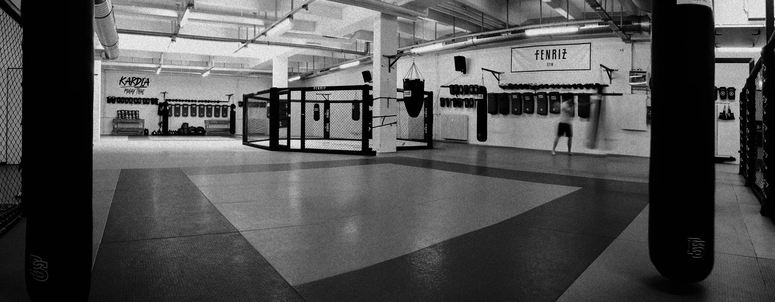 A black and white photograph of the gym with a man hitting the punching bag next to the cage.
